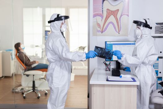 Dental receptionist dressed in coverall face shiled giving doctor patient x-ray keeping social distancing during covid19 virus pandemic. Woman waiting doctor diagnosis.