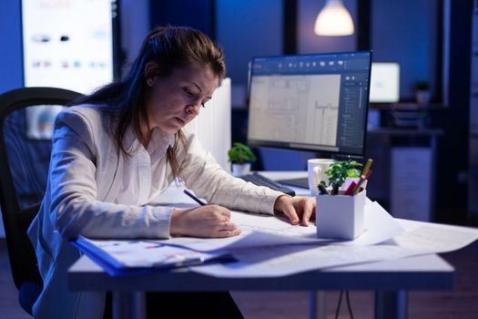Overworked woman architect checking and matching blueprints sitting at office desk
