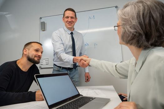 The boss makes a presentation to subordinates at the white board. Caucasian man shaking hands with middle aged woman.