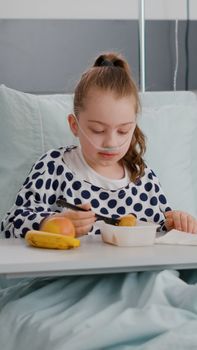 Portrait of hospitalized little child resting in bed eating healthy food meal during recovery examination