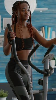 Black athletic woman doing cardio training on elliptical bike in living room