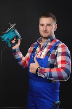 Smiling man in a plaid shirt and overalls holds a jigsaw in his hand on a black background and with the other hand shows a thumbs up gesture