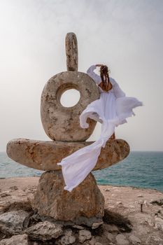 A woman stands on a stone sculpture made of large stones. She is dressed in a white long dress, against the backdrop of the sea and sky. The dress develops in the wind