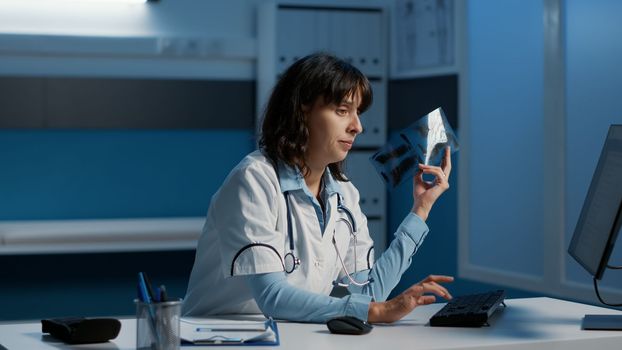 Practitioner holding radiography analyzing lungs before writing medical expertise on computer