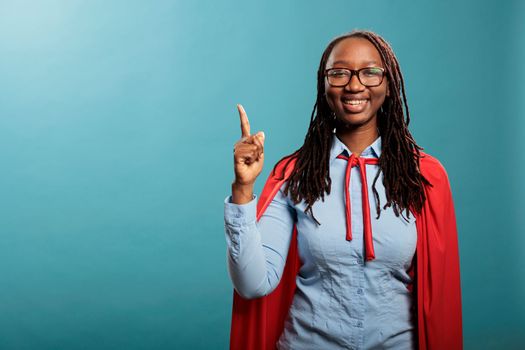 Optimistic happy superhero woman wearing red cape pointing finger up