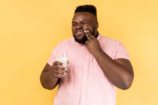 Portrait of sick sad upset man wearing pink shirt holding glass of water with ice, feels pain, touching his cheek, has sensitive teeth. Indoor studio shot isolated on yellow background.