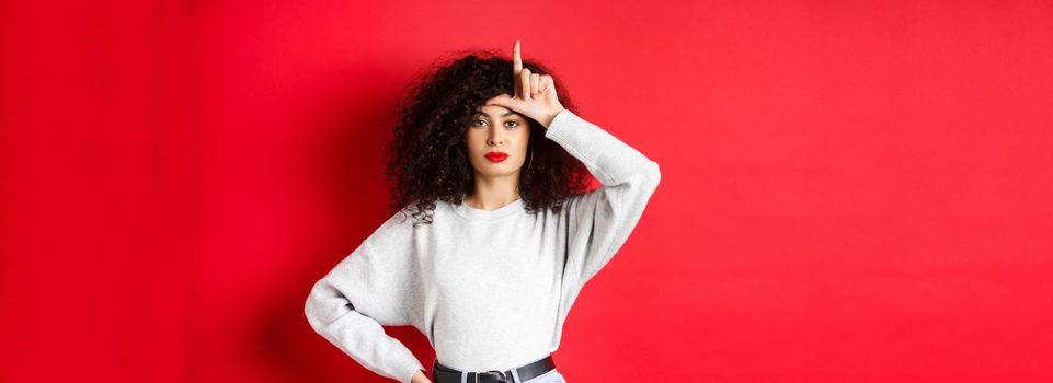 Confident european woman with curly hair, mocking lost team, showing loser gesture and staring unimpressed at camera, standing in casual clothes on red background