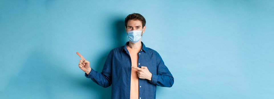 Covid and healthcare concept. Handsome modern guy in medical mask pointing at empty space aside, standing on blue background