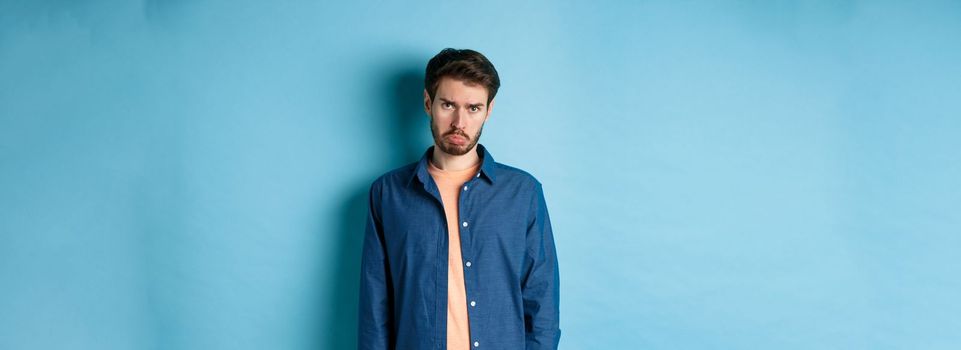 Sad and gloomy young man sulking, pulling miserable face and looking pleading at camera, standing on blue background