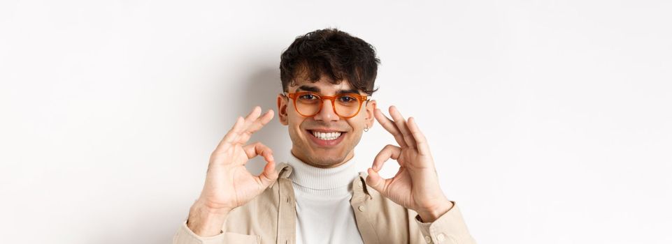 Close-up of handsome guy in new eyewear showing okay signs and smiling pleased, praising good glasses, standing on white background