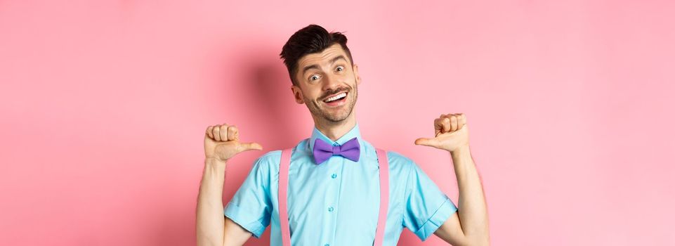 Cheerful smiling man with moustache and bow-tie, pointing at himself and self-promoting, standing happy over pink background in suspenders