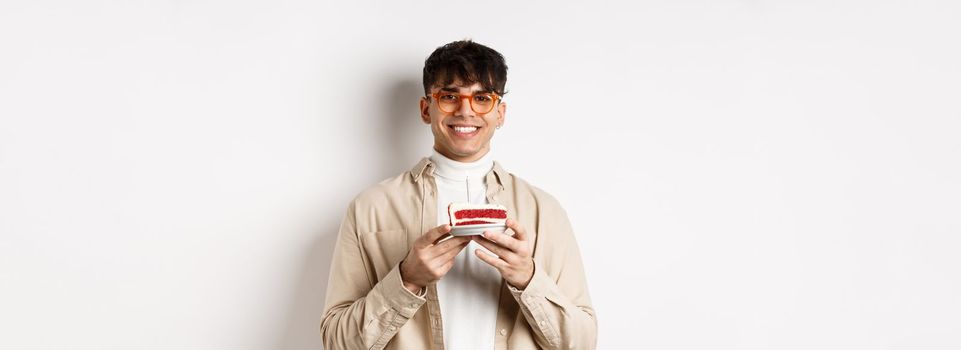Real people and holidays concept. Happy hipster guy in glasses celebrating birthday, making wish on b-day cake with candle and smiling, standing on white background