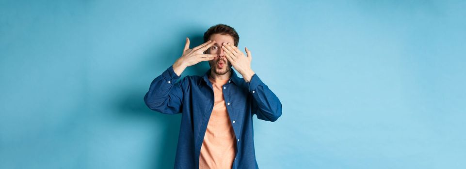 Excited young man peeking through fingers and saying wow, checking out surprise gift, standing on blue background