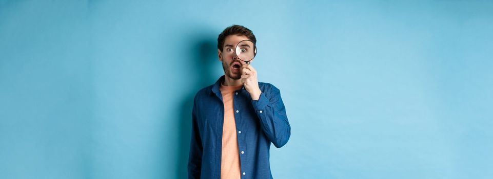 Impressed man look in awe through magnifying glass, drop jaw and stare at camera, standing on blue background