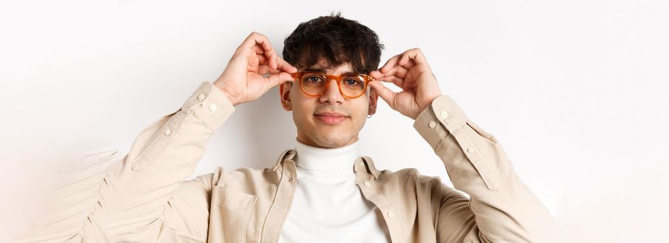 Close-up of stylish hipster guy trying eyewear at optician store, put on glasses and smiling, standing on white background