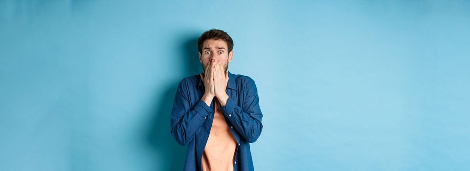 Scared anxious man covering mouth with hands and looking at something terrifying, standing on blue background
