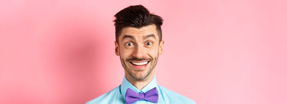 Close-up of cheerful caucasian man smiling happy, looking at something interesting, standing in bow-tie over pink background