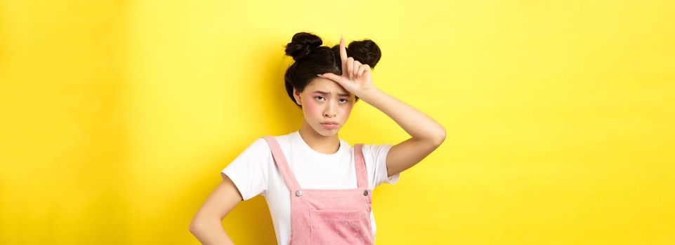 Sad girl showing loser sign on forehead and sulking upset, feeling disappointed in herself, standing on yellow background