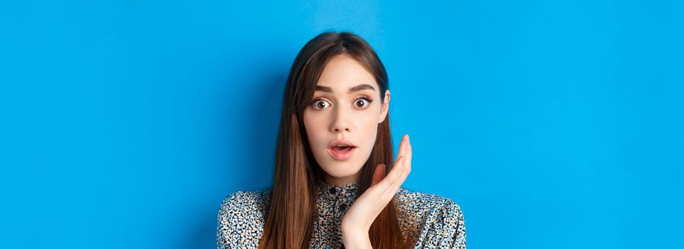Candid girl with long hair and makeup, looking shocked and surprised, open mouth and stare speechless at camera, standing on blue background