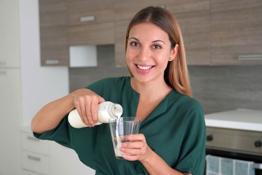 Morning portrait of beautiful smiling woman filling the glass with kefir. Looks at camera.