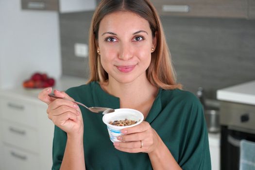 Attractive woman holds bowl of Greek yogurt with granola and smiles at camera with blurred kitchen on the background