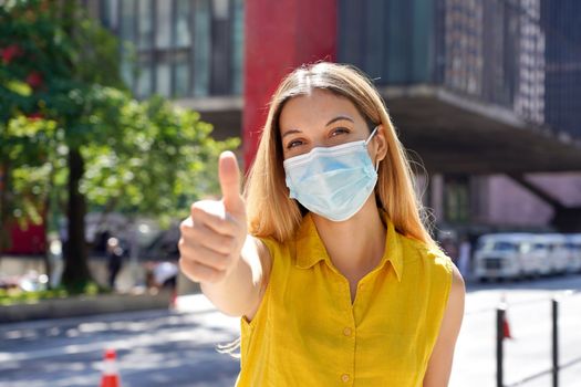 Optimistic young woman wearing protective surgical mask showing thumbs up in Sao Paulo city street, Brazil