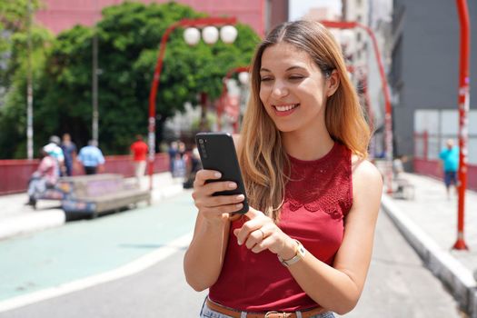 Portrait of smiling Brazilian woman walking in Sao Paulo City typing on smartphone with blurred background