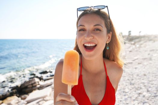 Beautiful excited woman laughing with a popsicle in her hand on the beach on summer. Looks at camera.