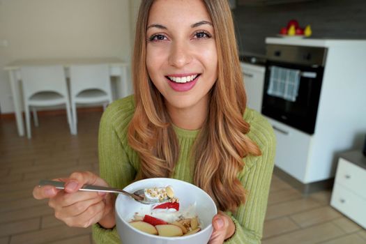Young woman eating muesli granola oatmeal with fruits and yogurt looking at camera in kitchen