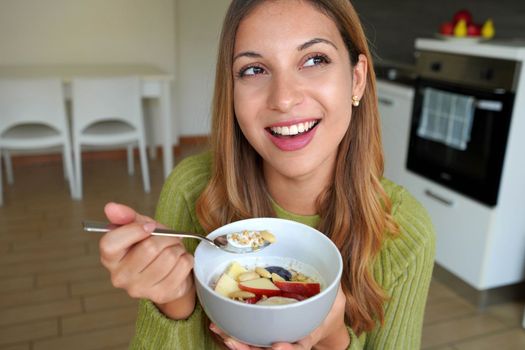 Beautiful woman eating muesli granola oatmeal with fruits and yogurt looking to the side at home
