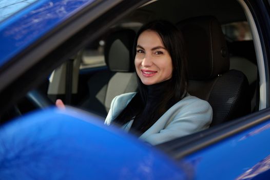 Attractive middle-aged European woman in formal wear, driving car and smiling a beautiful toothy smile looking at camera