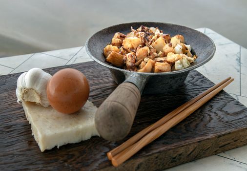 Traditional stir fried soft turnip cake or Fried radish cake (chai tow kway) in small steaming iron pot Served with wooden chopsticks on white table. 
