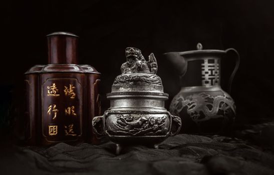 Silver antique incense burner, Chinese antique tea leaf iron storage jar (Characters chinese is Name of the tea) and Chinese antique teapot (Characters chinese is Double Happiness) on dark background. 