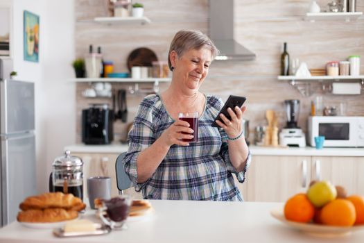 Senior woman using mobile gadget in the kitchen drinking aromatic tea