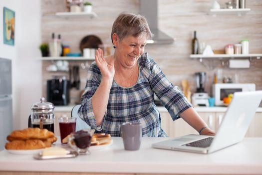 Retired woman waving during online meeting with family on video call using laptop
