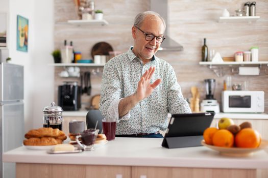 Mature man waving while having a conversation during video call in kitchen