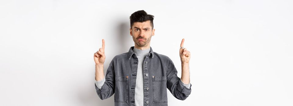 Confused funny guy with moustache pointing fingers up at something strange, frowning and pouting puzzled, standing on white background