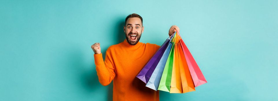 Happy handsome man holding shopping bags and smiling, feeling excitement from discounts, making fist pump, standing over turquoise background