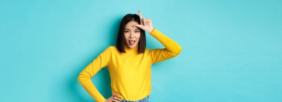 Asian girl winner mocking over lost team, showing loser sign on forehead and sticking tongue ignorant, standing over blue background