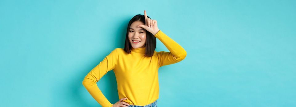 Sassy asian girl mocking lost team, showing loser sign on forehead and smiling pleased, being a winner, standing over blue background