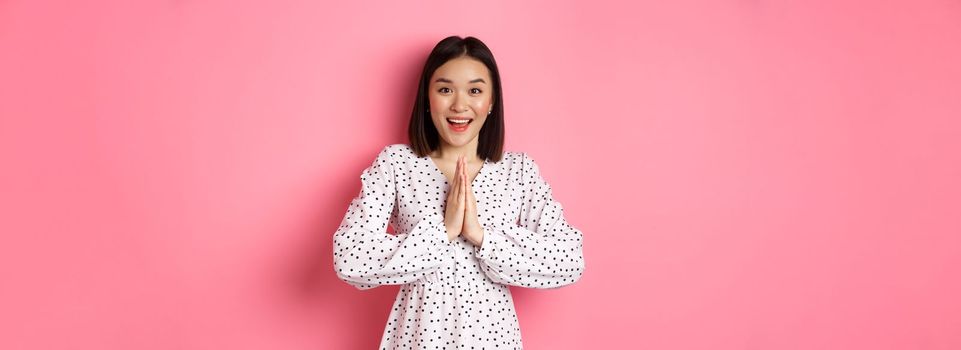 Beautiful asian woman thanking you, holding hands together in appreciation gesture, smiling happy at camera, standing grateful over pink background