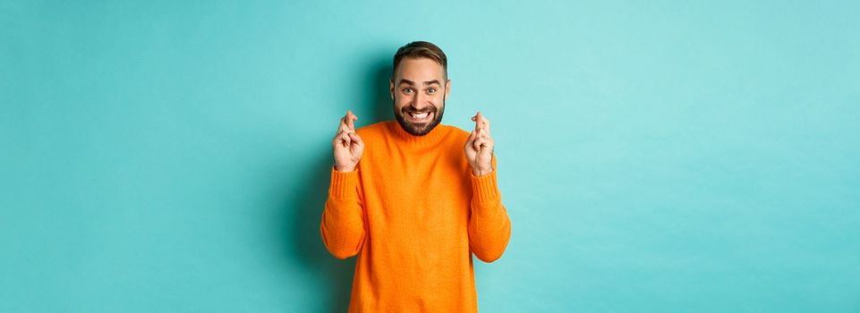 Excited man with beard, making a wish, holding fingers crossed for good luck and smiling, standing over light blue background