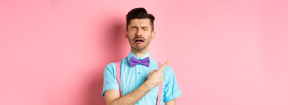 Sad and lonely man in bow-tie crying and pointing finger left, feeling miserable and complaining, sulking while standing on pink background