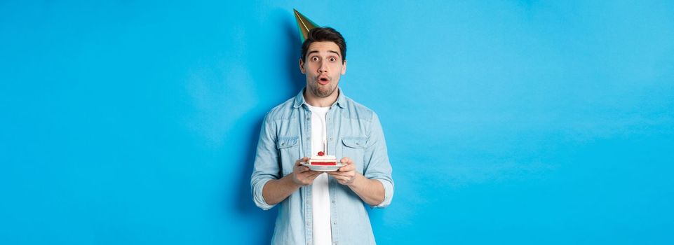 Handsome man in party cone holding birthday cake, looking surprised, standing over blue background