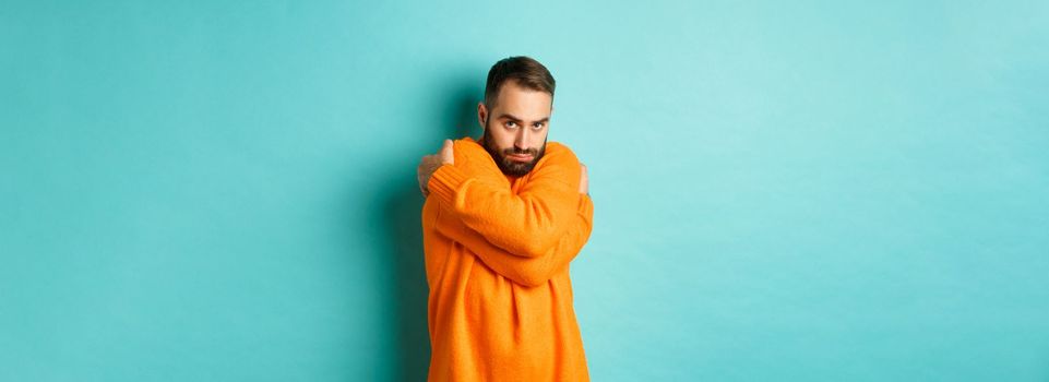 Timid man feeling offended and defensive, hugging himself and looking suspicious at camera, standing over light blue background
