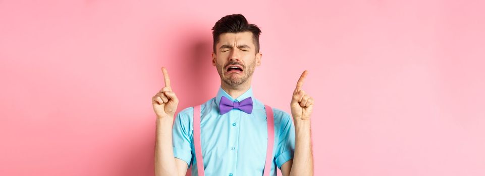 Sad and miserable guy sobbing and crying, pointing fingers up and something disappointed, standing upset on pink background
