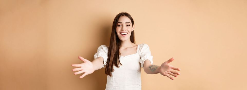 Portrait of friendly candid girl reaching for hug, welcome friend and smiling, spread hands sideways in greeting gesture, receiving something, beige background