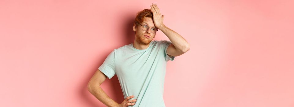 Tired young man with red hair, wearing glasses, looking annoyed and tensed, making facepalm gesture and exhale bothered, standing over pink background