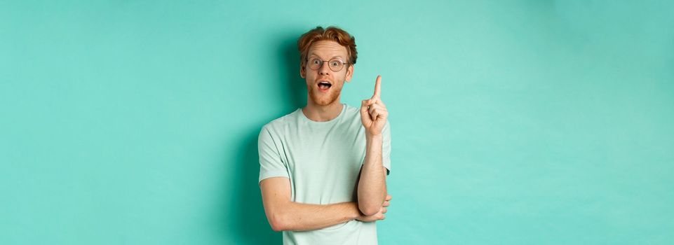 Excited young man with ginger hair in glasses, raising index finger, pitching an idea, standing over mint background