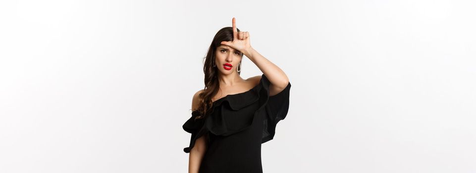 Fashion and beauty. Arrogant glamour woman showing loser sign on forehead, mocking lost person, standing in black dress over white background